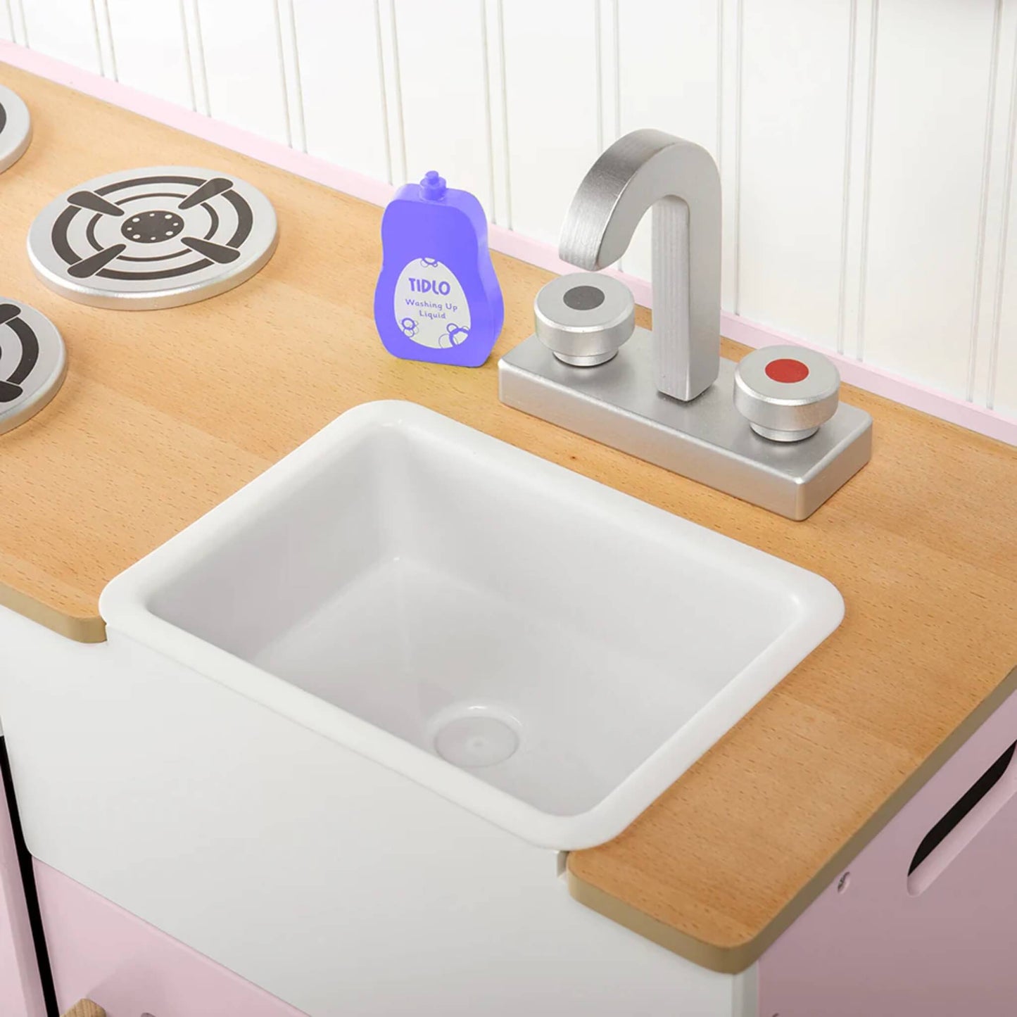 Close-up of the white plastic sink with silver taps and a bottle of pretend Tidlo washing-up liquid on a wooden counter beside four hob plates.