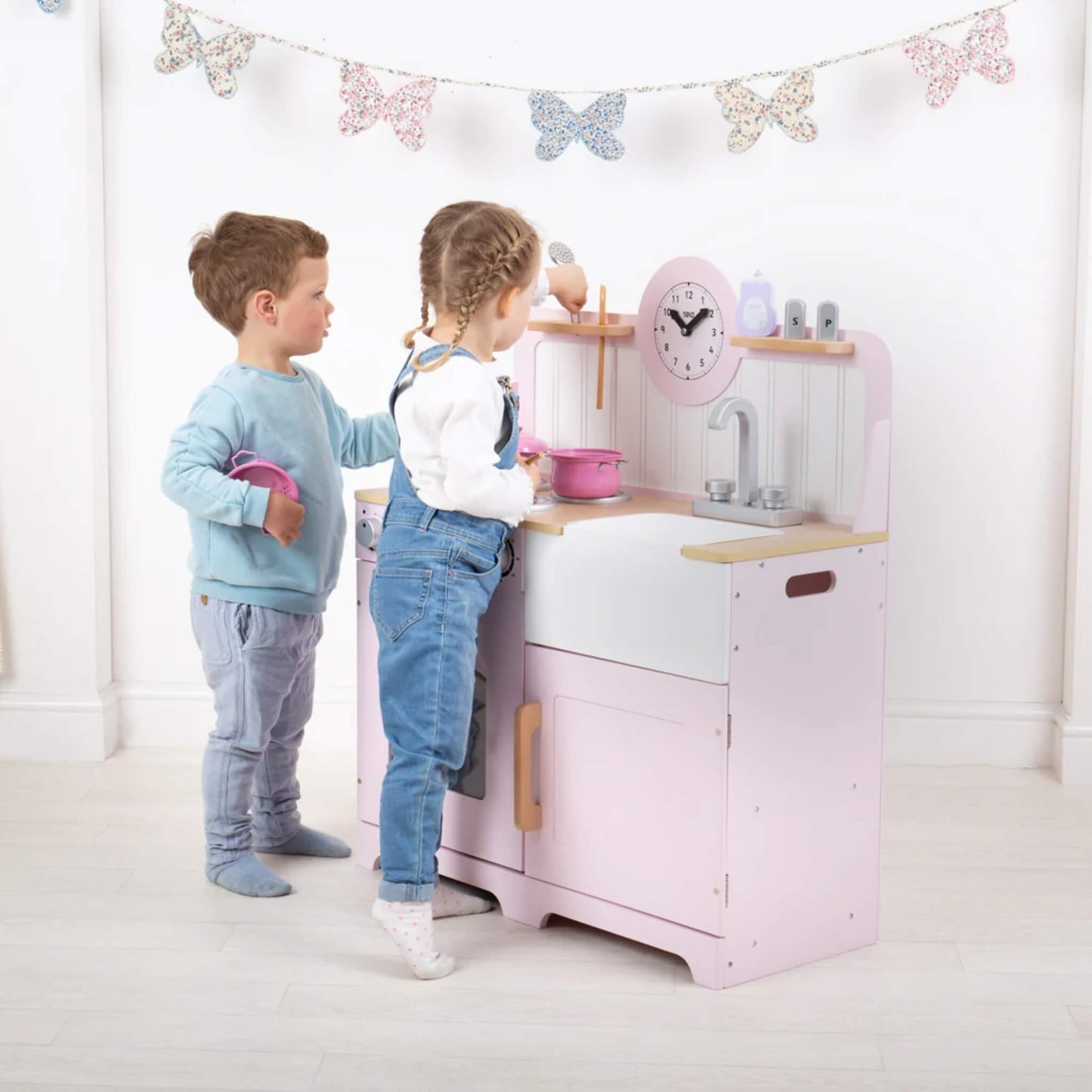 Two children play side by side at the pink Tidlo wooden kitchen, pretending to cook with a saucepan and utensils under a string of decorative butterfly bunting.