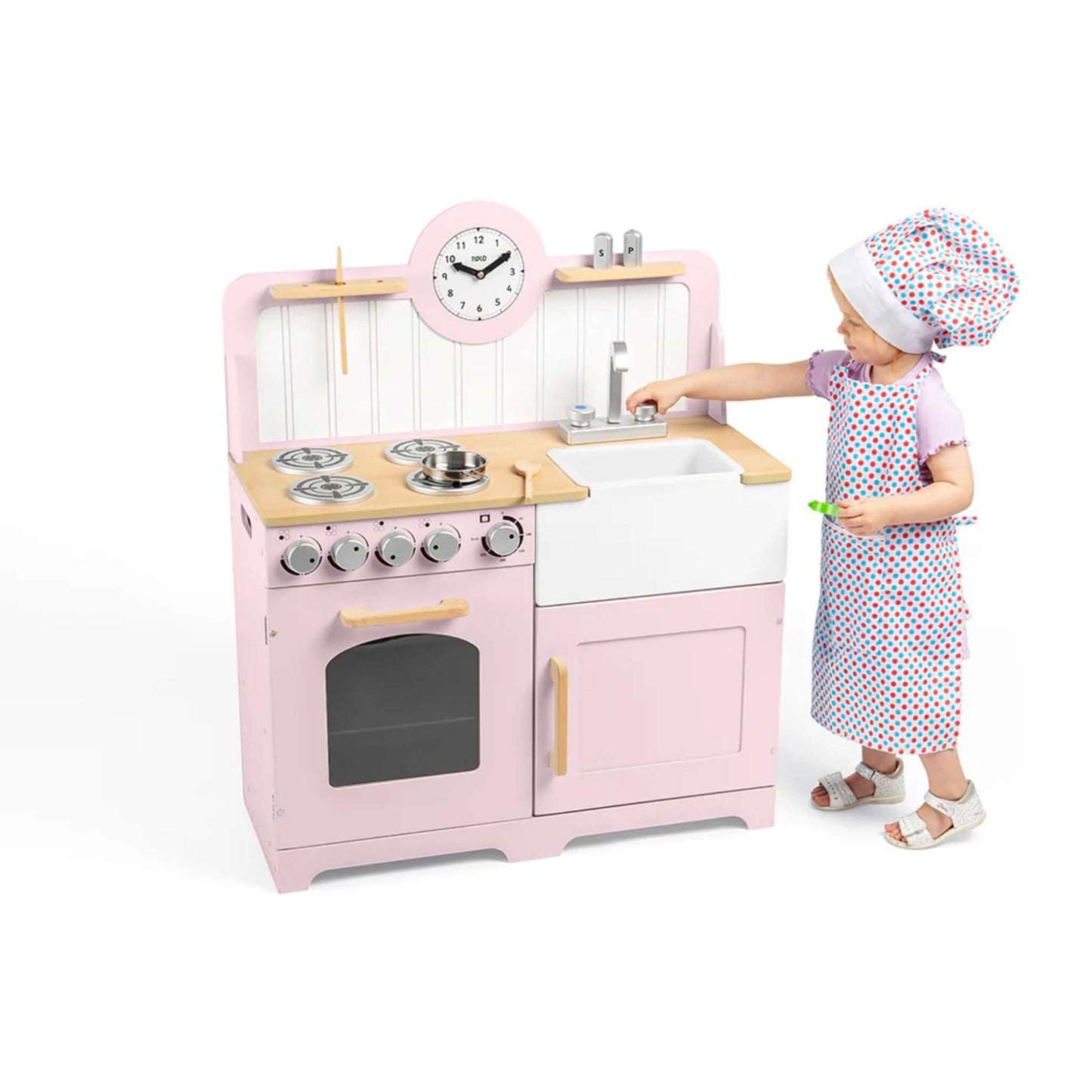 A toddler in an apron and chef’s hat pretends to wash up at the pink wooden play kitchen, turning the tap above the Belfast sink.