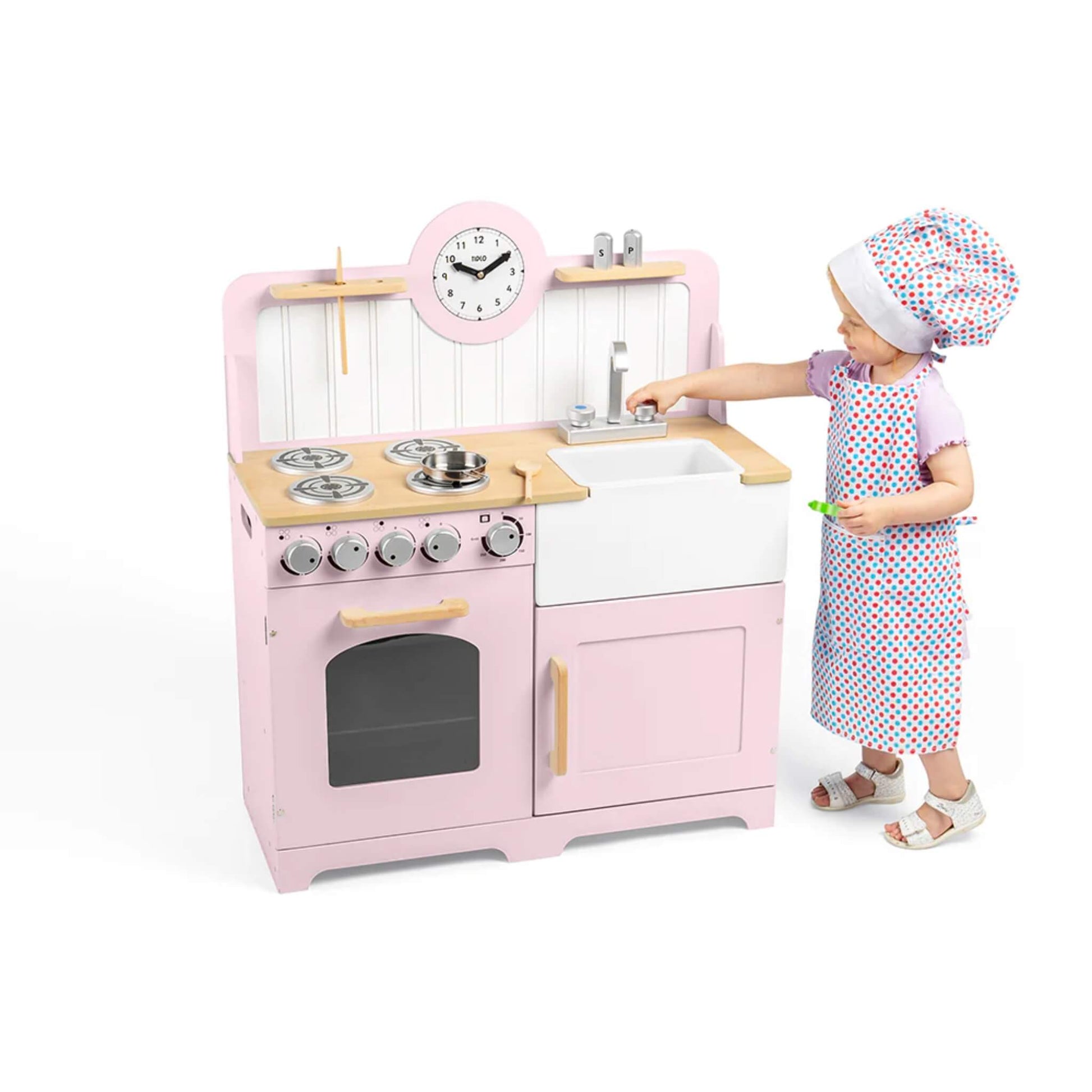 A toddler in an apron and chef’s hat pretends to wash up at the pink wooden play kitchen, turning the tap above the Belfast sink.