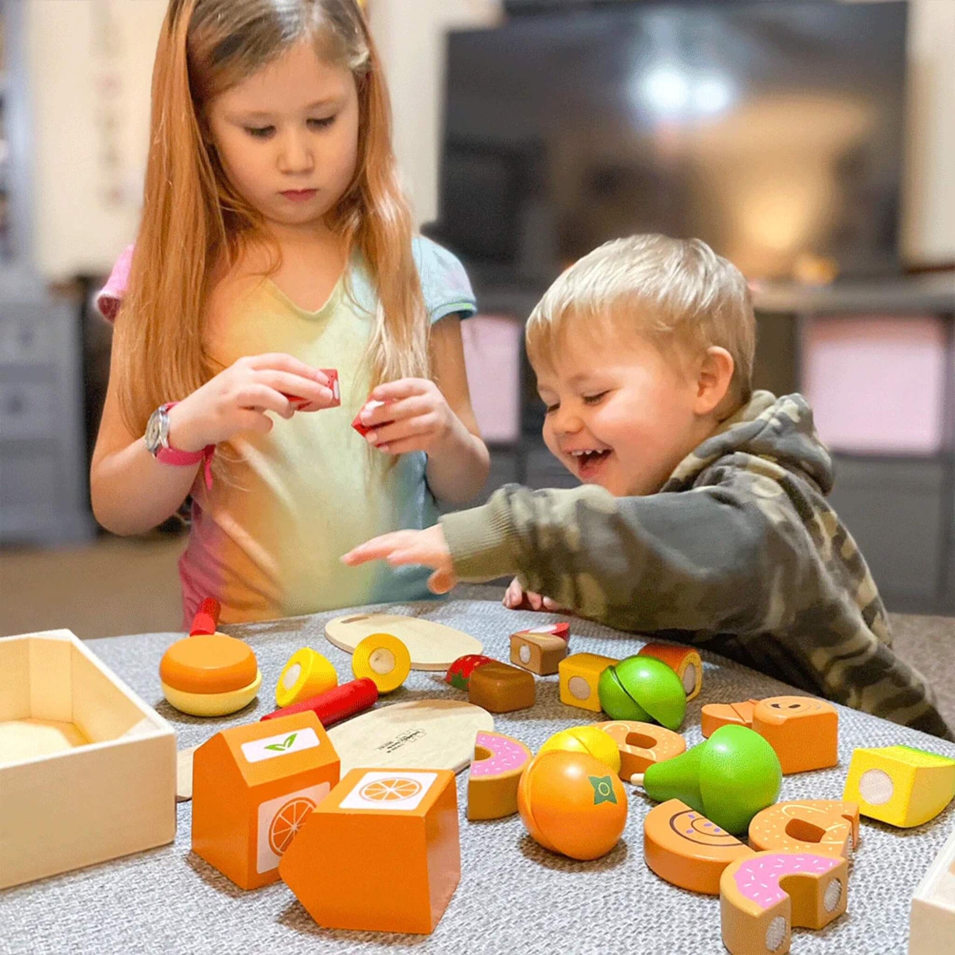 Two children playing at a table with an assortment of wooden play food pieces, including fruit slices, burger parts and boxed grocery items.