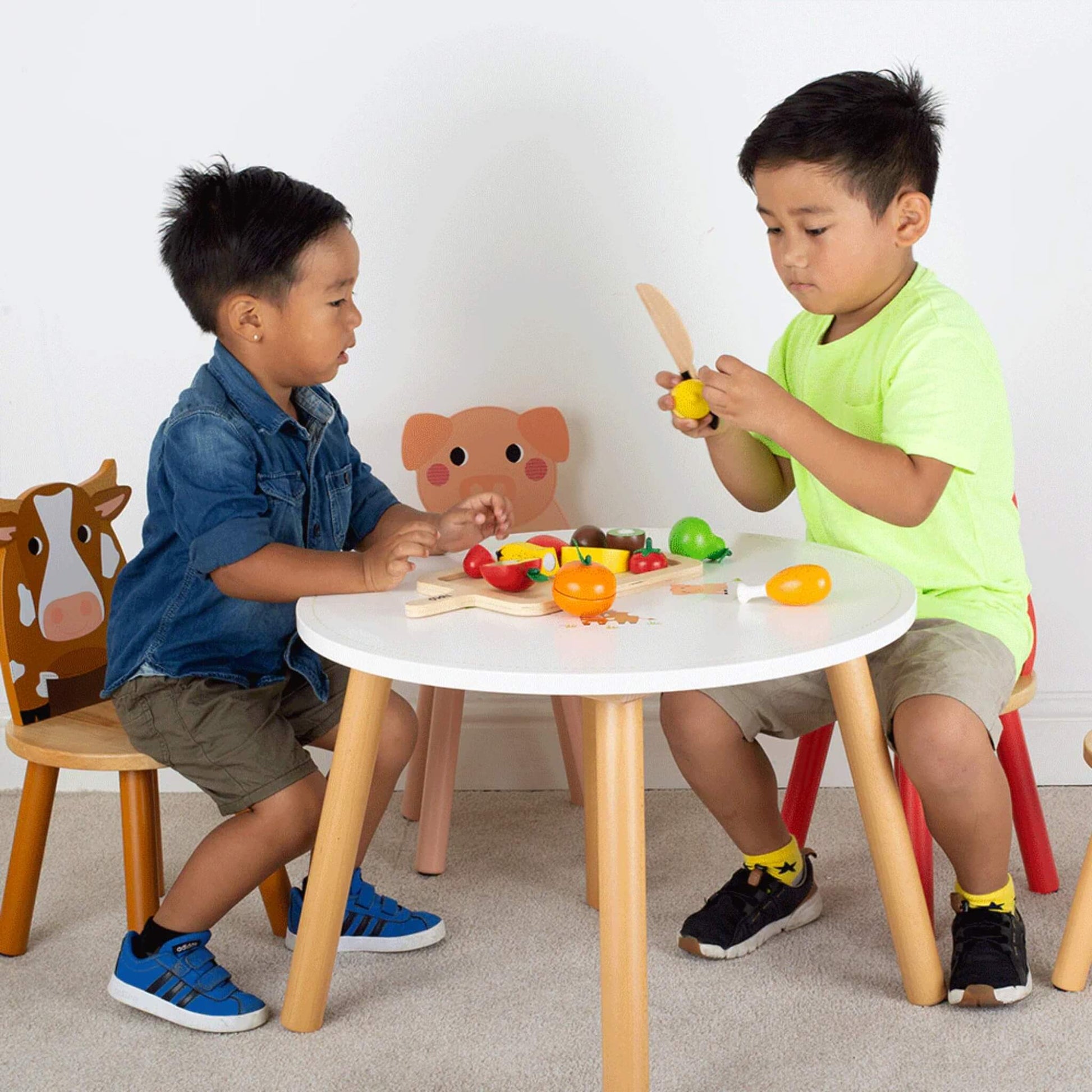 Two young boys sit at a small white table cutting and arranging wooden fruit pieces from the Tidlo set, with a chopping board and tray between them.