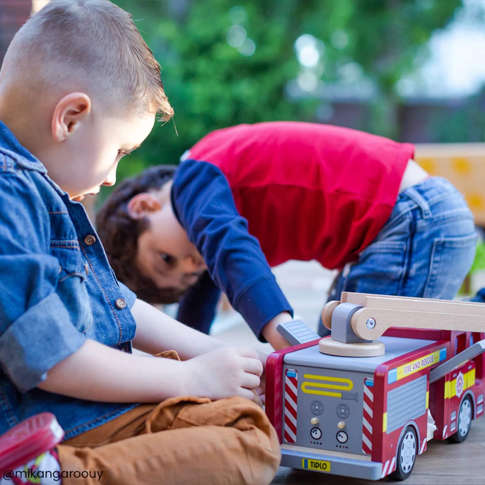 Two young boys crouched on a patio playing with the Tidlo Fire Engine, shown from the back with a visible rotating ladder.