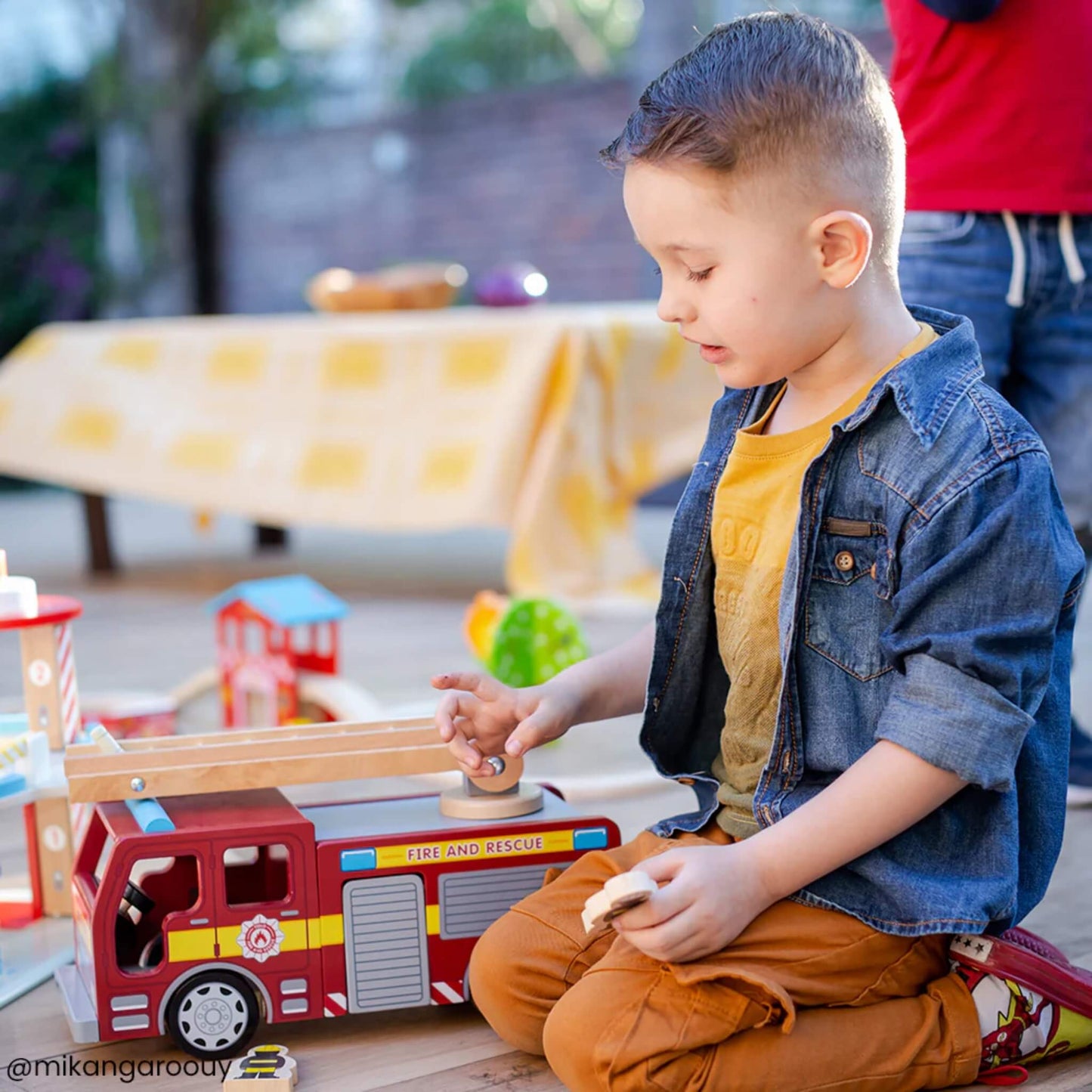 A young boy kneeling and playing with the Tidlo Fire Engine outdoors, shown smiling and interacting with the wooden accessories.