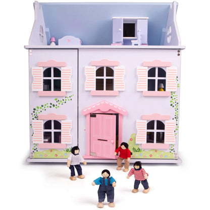 Wooden doll family placed in front of a pastel pink-and-purple dollhouse with shutter windows and floral details, ready to move in.