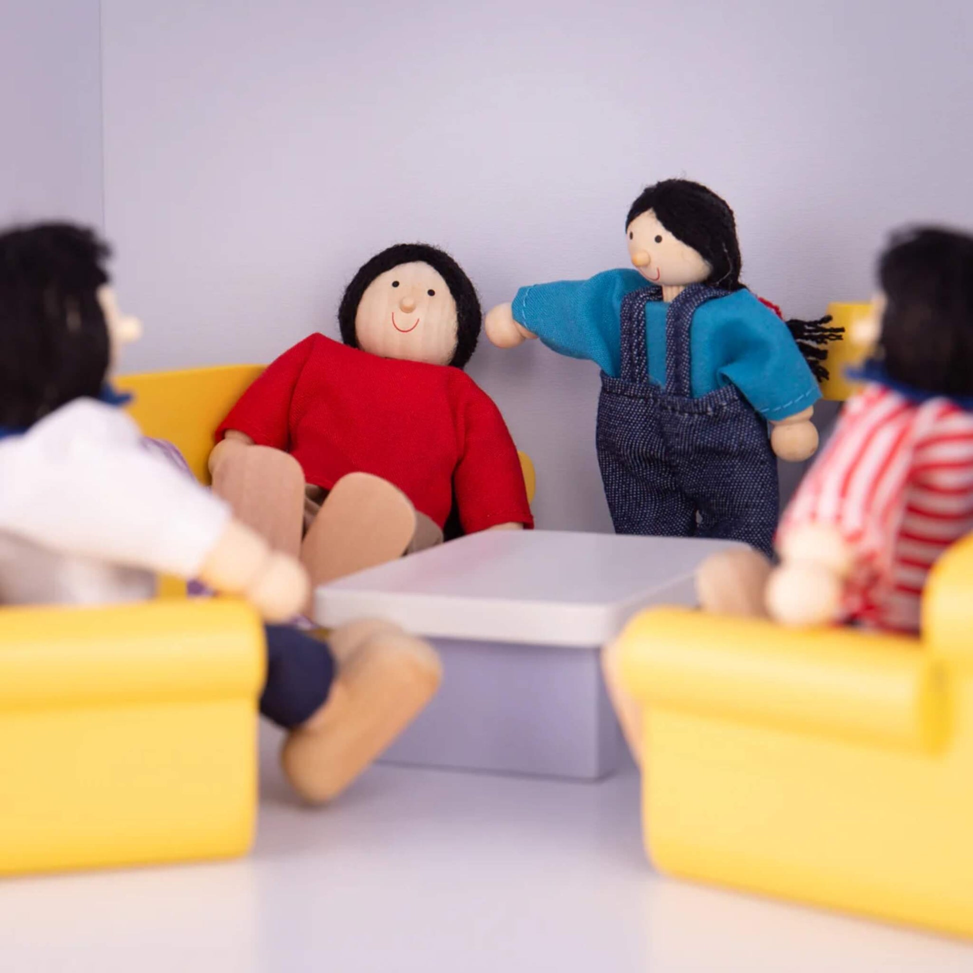 Close-up of the dolls arranged around a playroom table and yellow chairs, suggesting a family conversation or social moment.