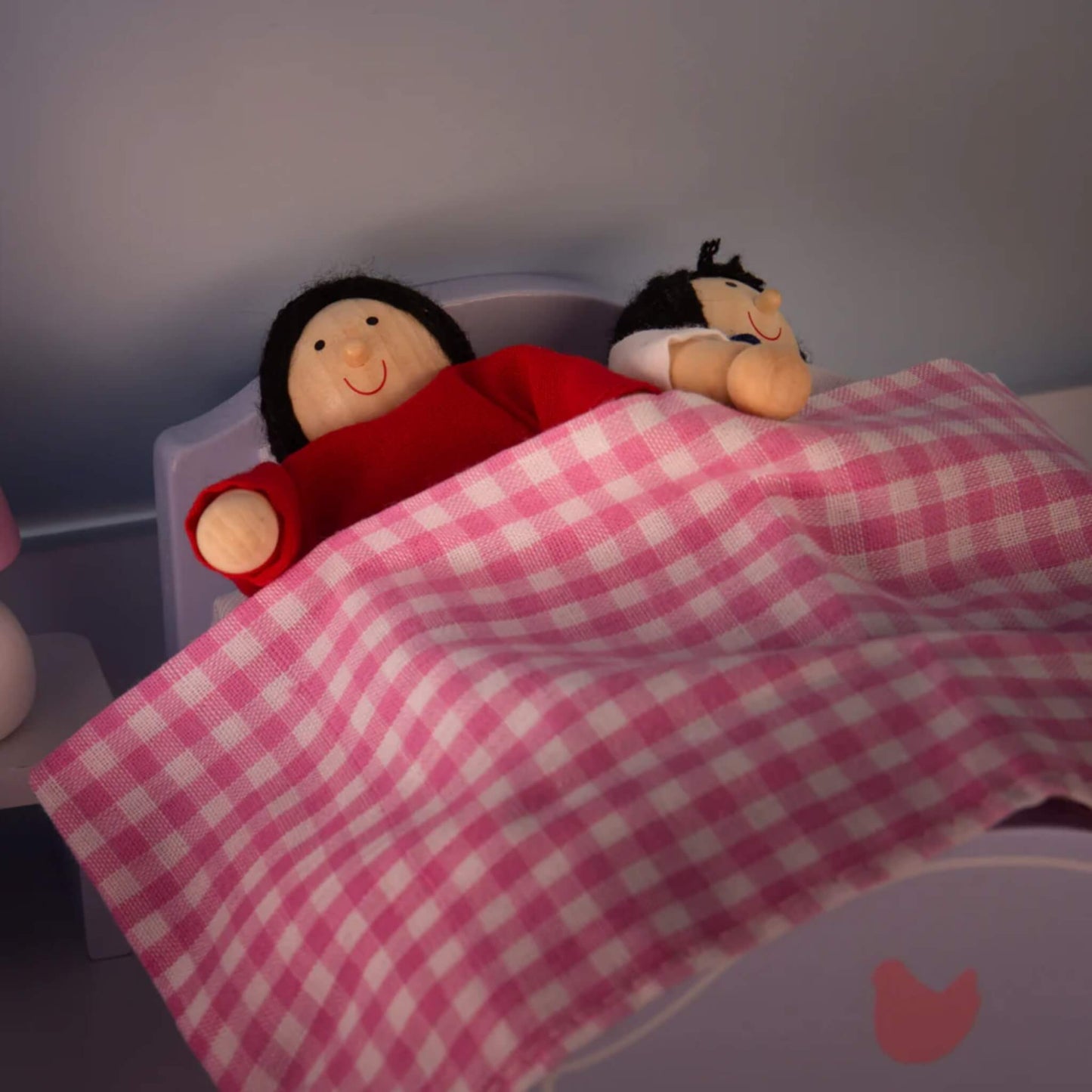 Two dolls tucked into a small bed with a pink gingham blanket, in a softly lit bedroom scene inside the dollhouse.