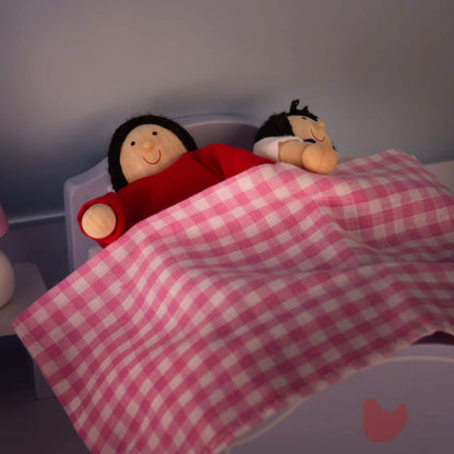 Two dolls tucked into a small bed with a pink gingham blanket, in a softly lit bedroom scene inside the dollhouse.