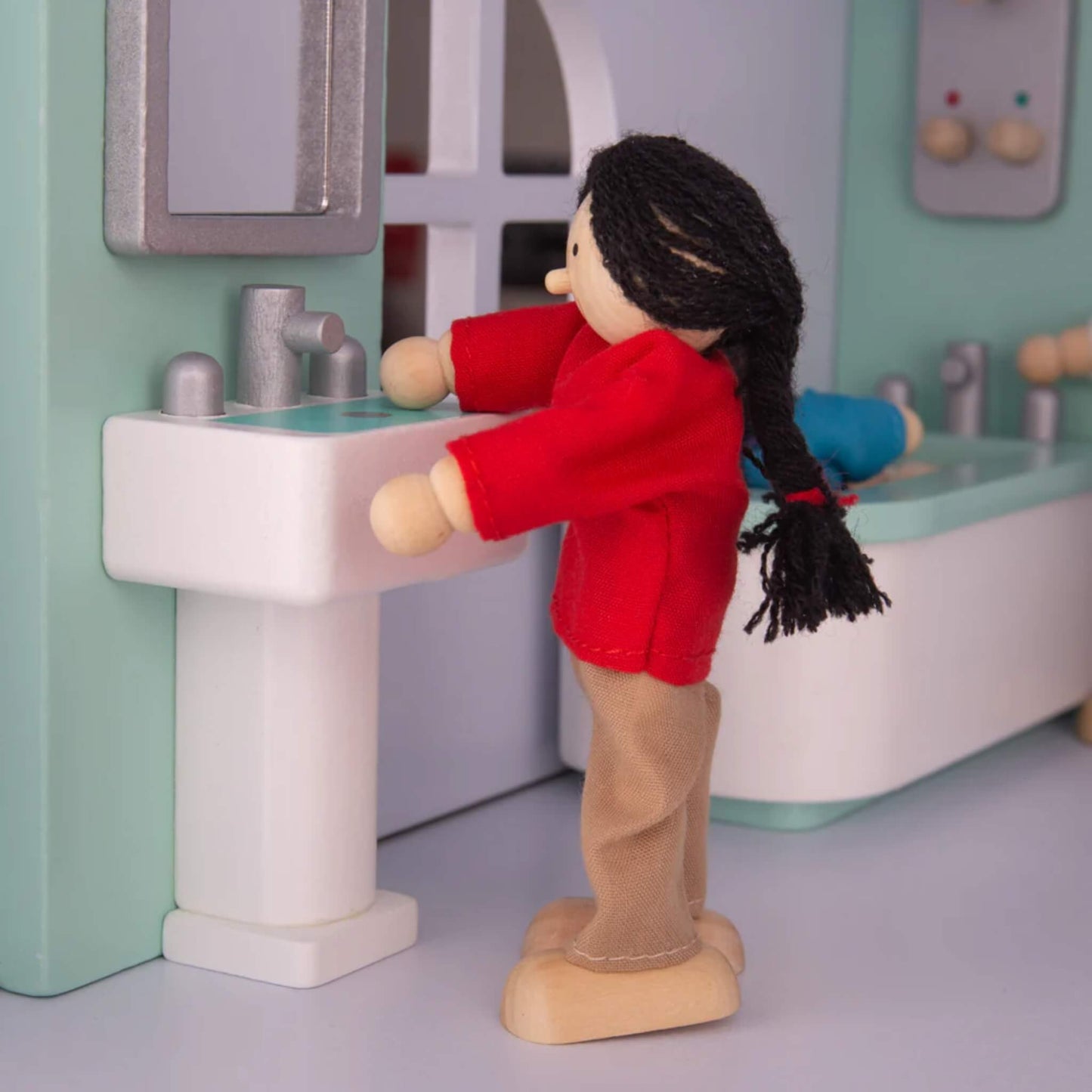 Side view of a doll in a red top standing at a miniature bathroom sink and mirror, mimicking handwashing or grooming.