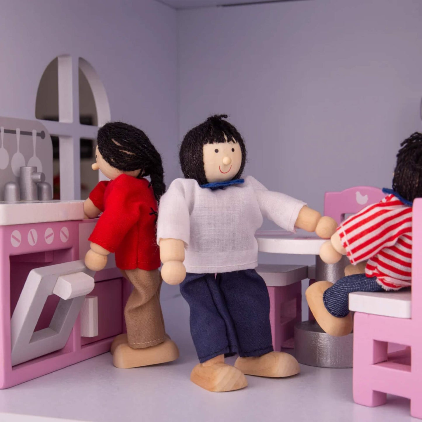 Dolls interacting in a kitchen and dining room area—one stands at the oven, while others are seated around a pink and white table.