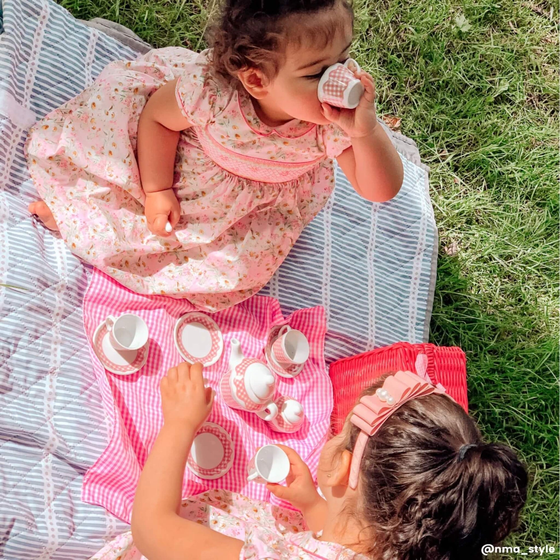 Two young children having a tea party on a picnic blanket, with gingham tea set items arranged in front of them, each child dressed in pink floral dresses.