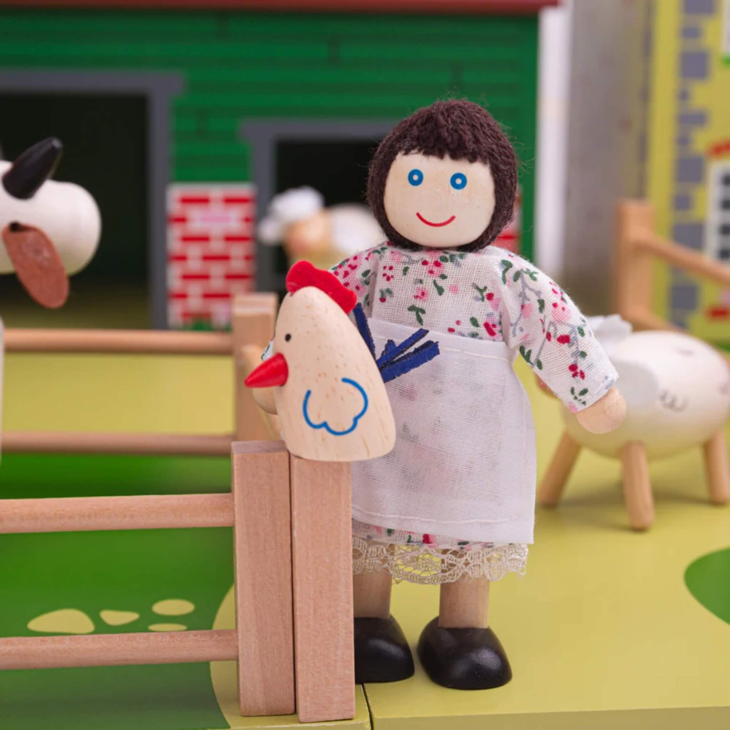 Smiling wooden farmer doll in a floral dress standing by the fence with a chicken from the Tidlo Oldfield Farm set, with animals grazing nearby.