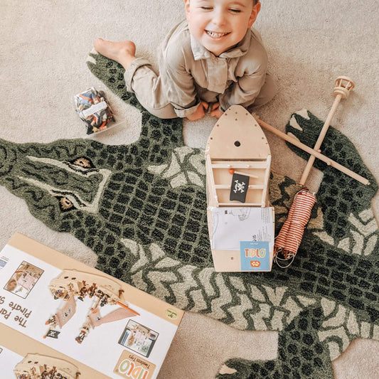 Young child smiling beside the unassembled Tidlo pirate ship with wooden parts and pirate figures laid out on a crocodile-shaped rug.
