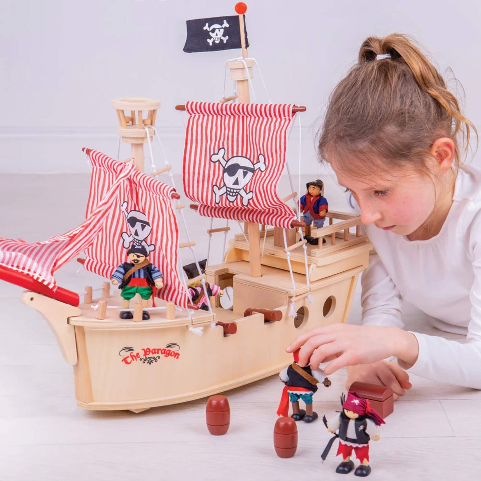Girl playing on the floor with Tidlo pirate ship, setting up figures and barrels around the wooden boat during imaginative play.