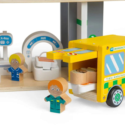 A children's wooden playset featuring a yellow ambulance with an open back, a white MRI scanner, an X-ray machine, and two small wooden character figures in blue and teal uniforms on a light wood base.