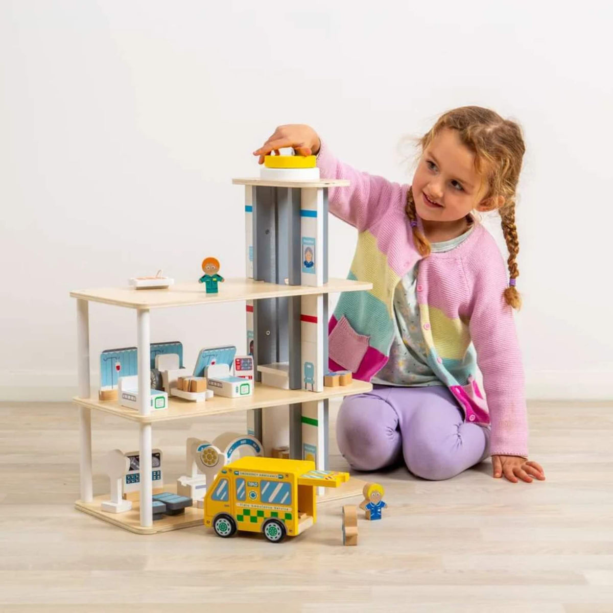A child playing with the wooden hospital, operating the lift with hospital beds, figures and yellow ambulance arranged across the floors.