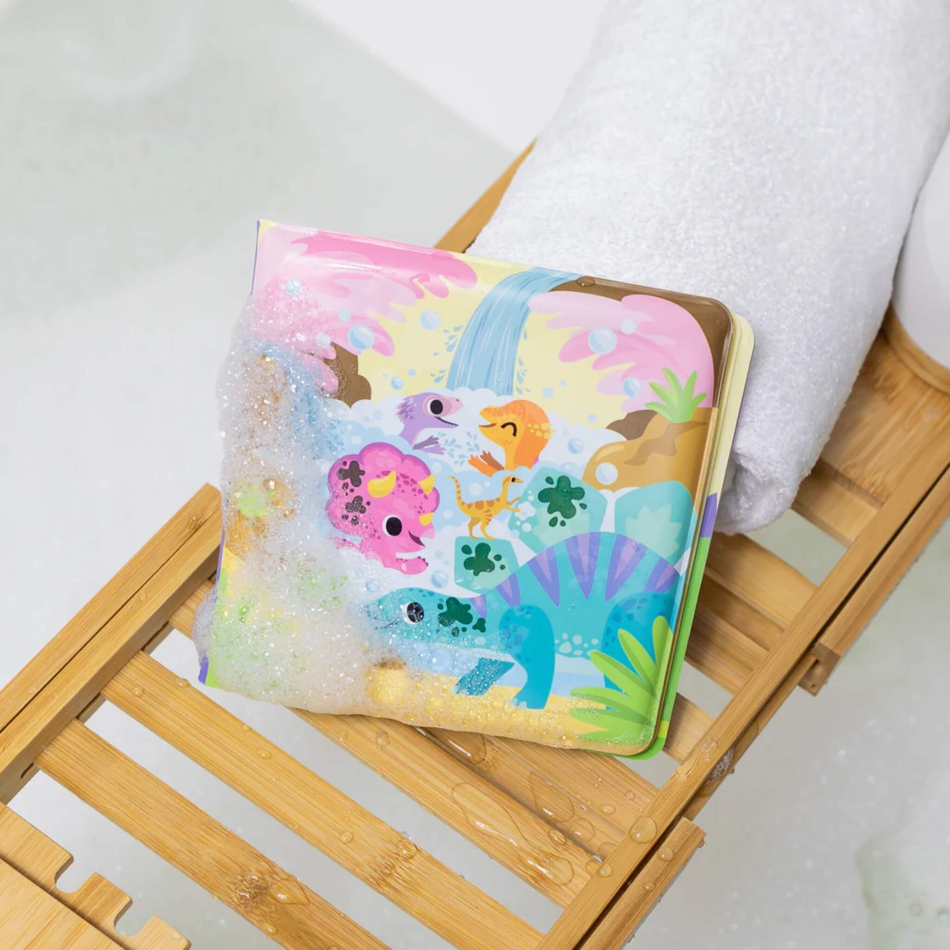 Tiger Tribe Messy Dinosaurs Bath Book resting on a wooden bath tray with bubbles, partially covered in foam and showing playful dinosaur illustrations.
