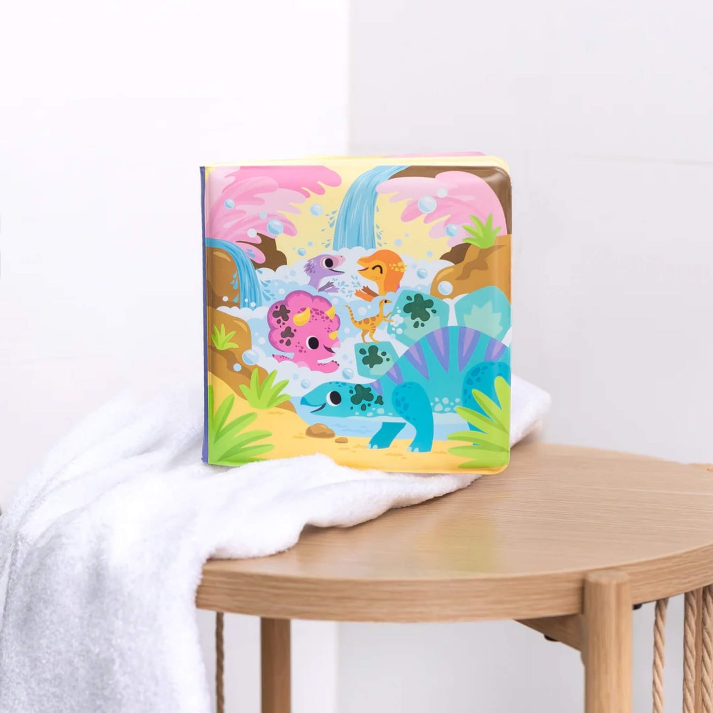 Bath book standing upright on a wooden stool, next to a fluffy white towel. Cover art with dinosaurs and waterfalls clearly visible.