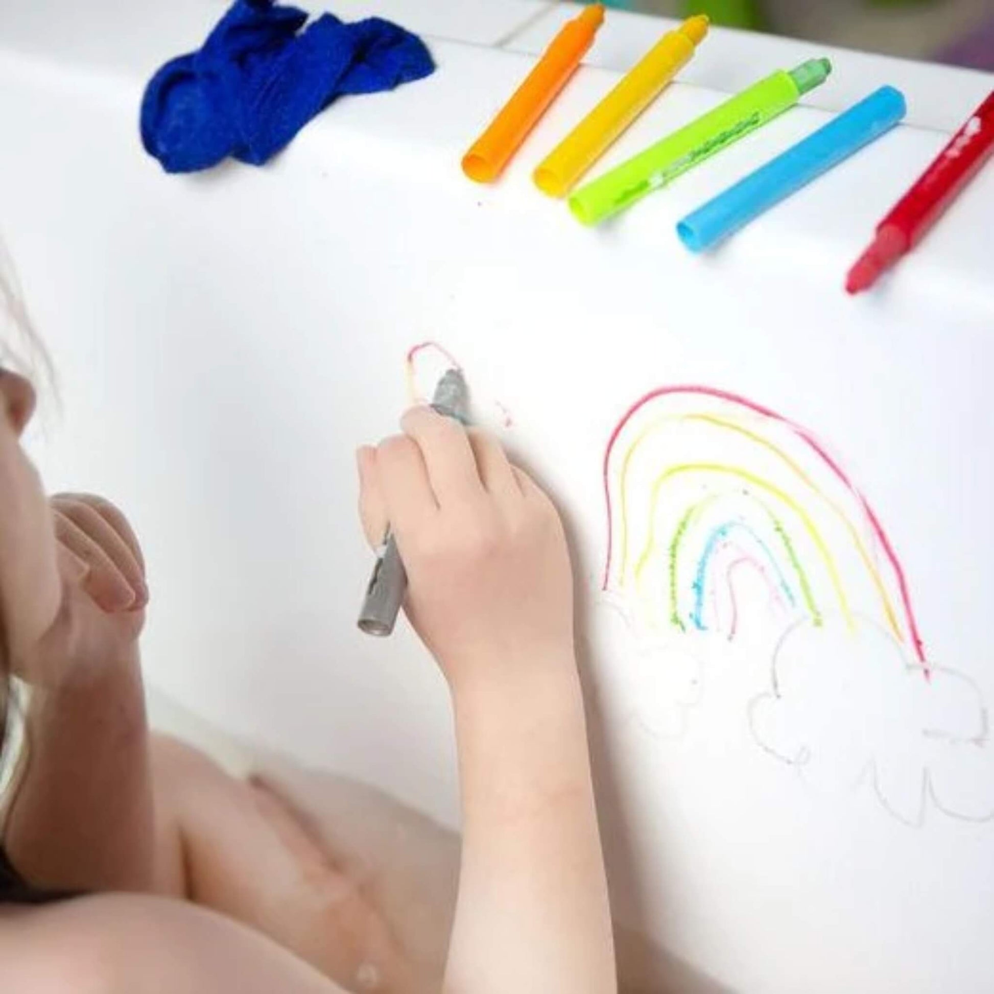A child in the bath draws a rainbow on the side of the tub using a grey Tiger Tribe bath crayon. Other crayons and a cloth are lined up on the edge.
