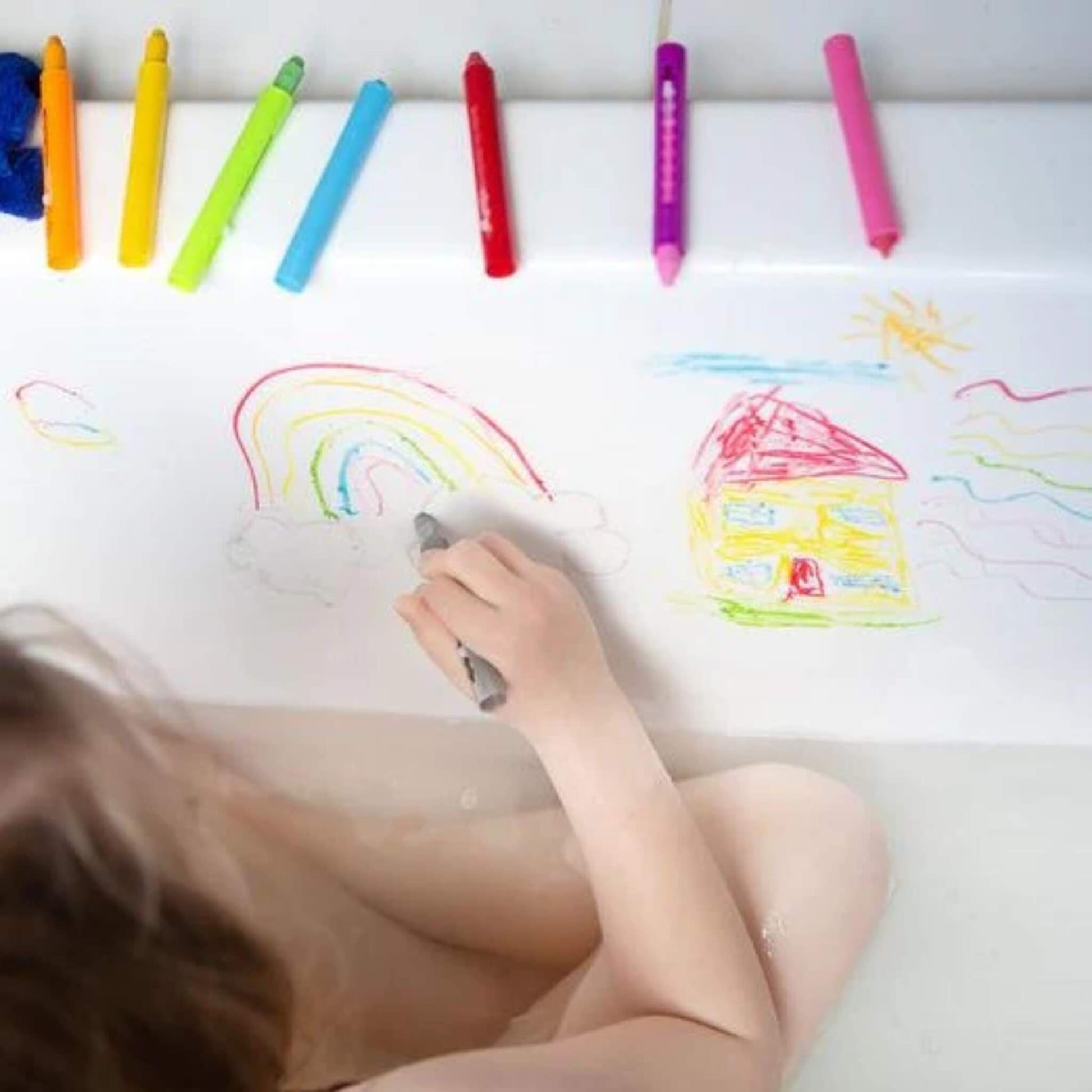 A child draws a colourful house and rainbow on the bath wall using Tiger Tribe bath crayons, with six open crayons resting nearby.