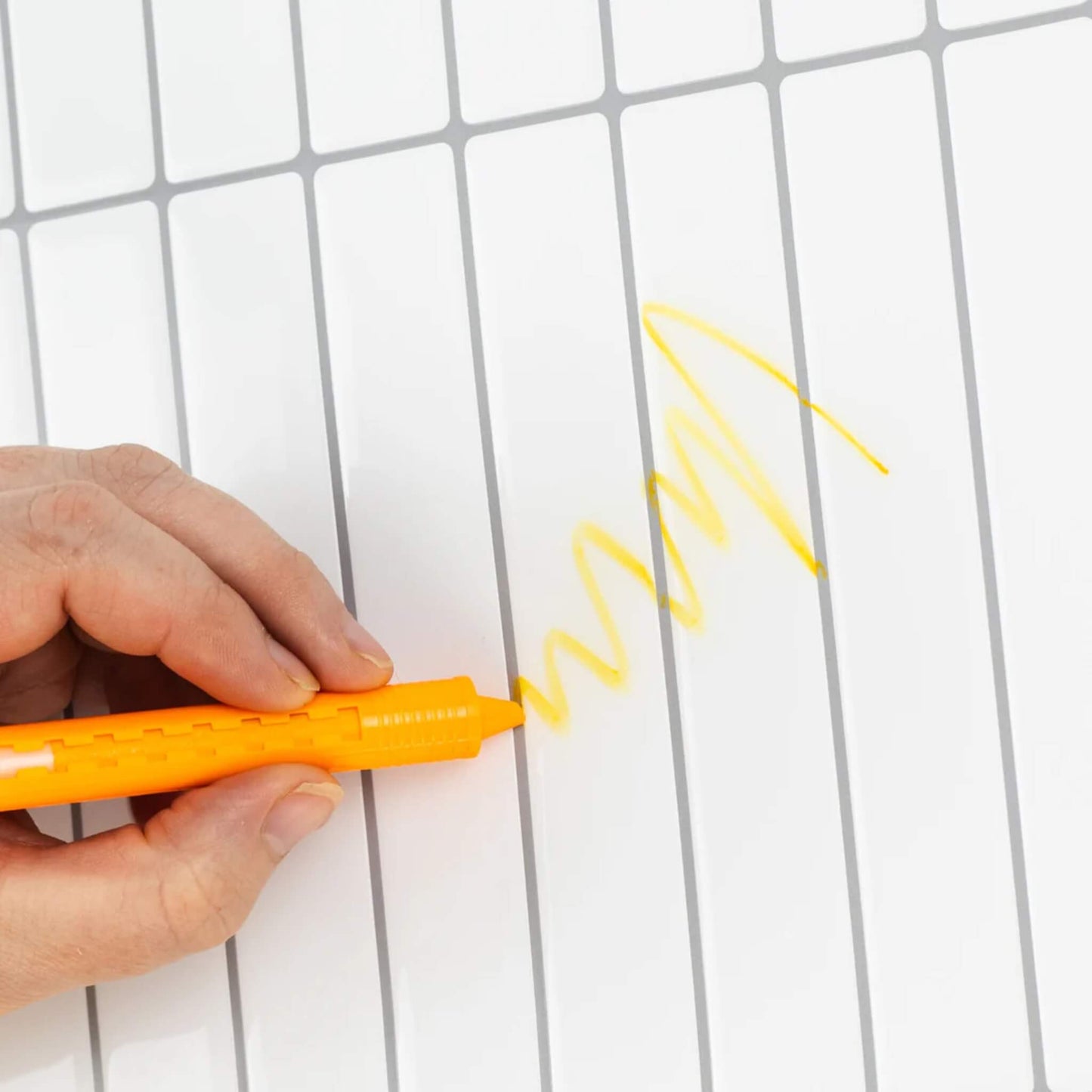 Close-up of a hand drawing a yellow squiggle on white bathroom tiles using a Tiger Tribe crayon.