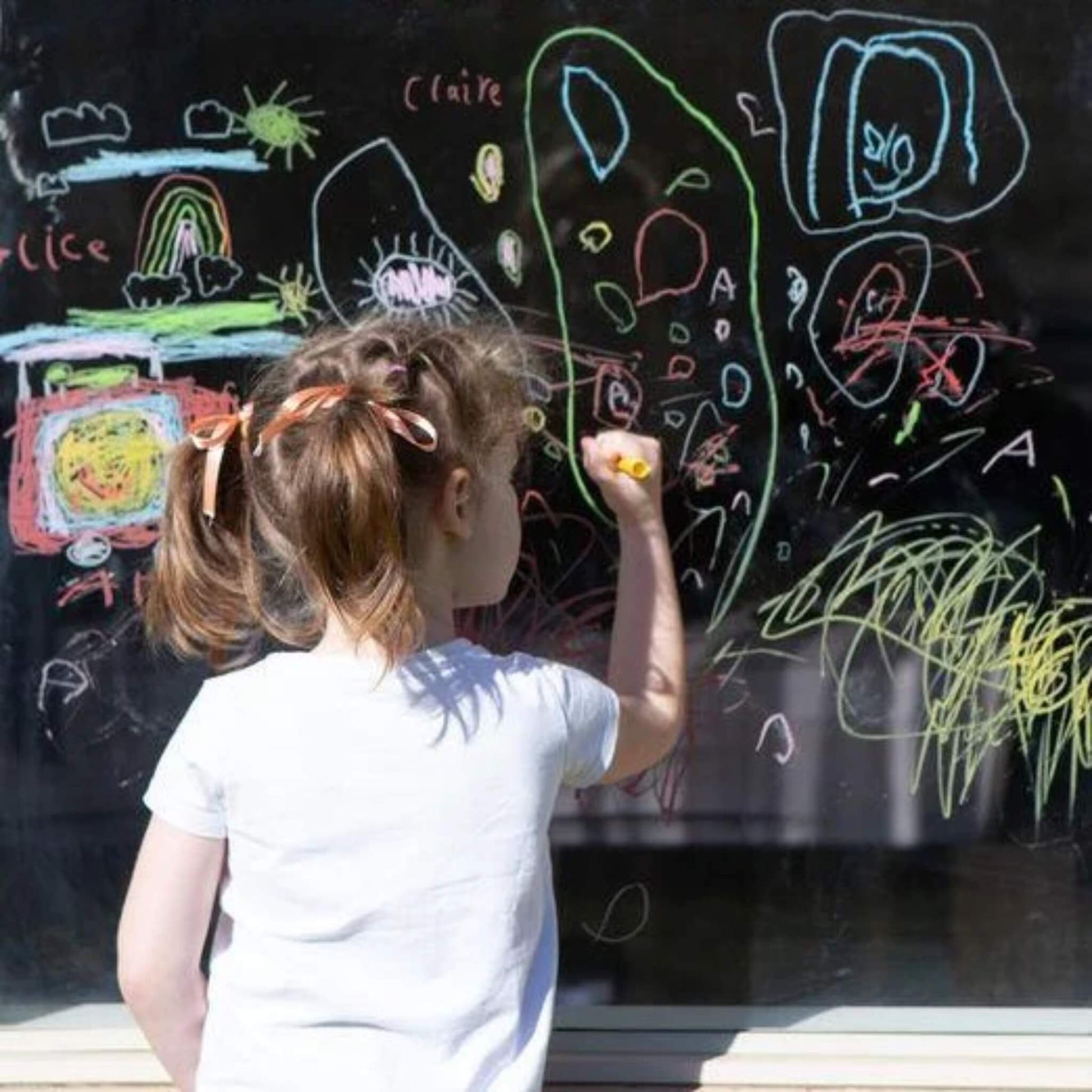 A girl draws on a window with colourful Tiger Tribe crayons, surrounded by drawings of rainbows, stars, and abstract shapes.