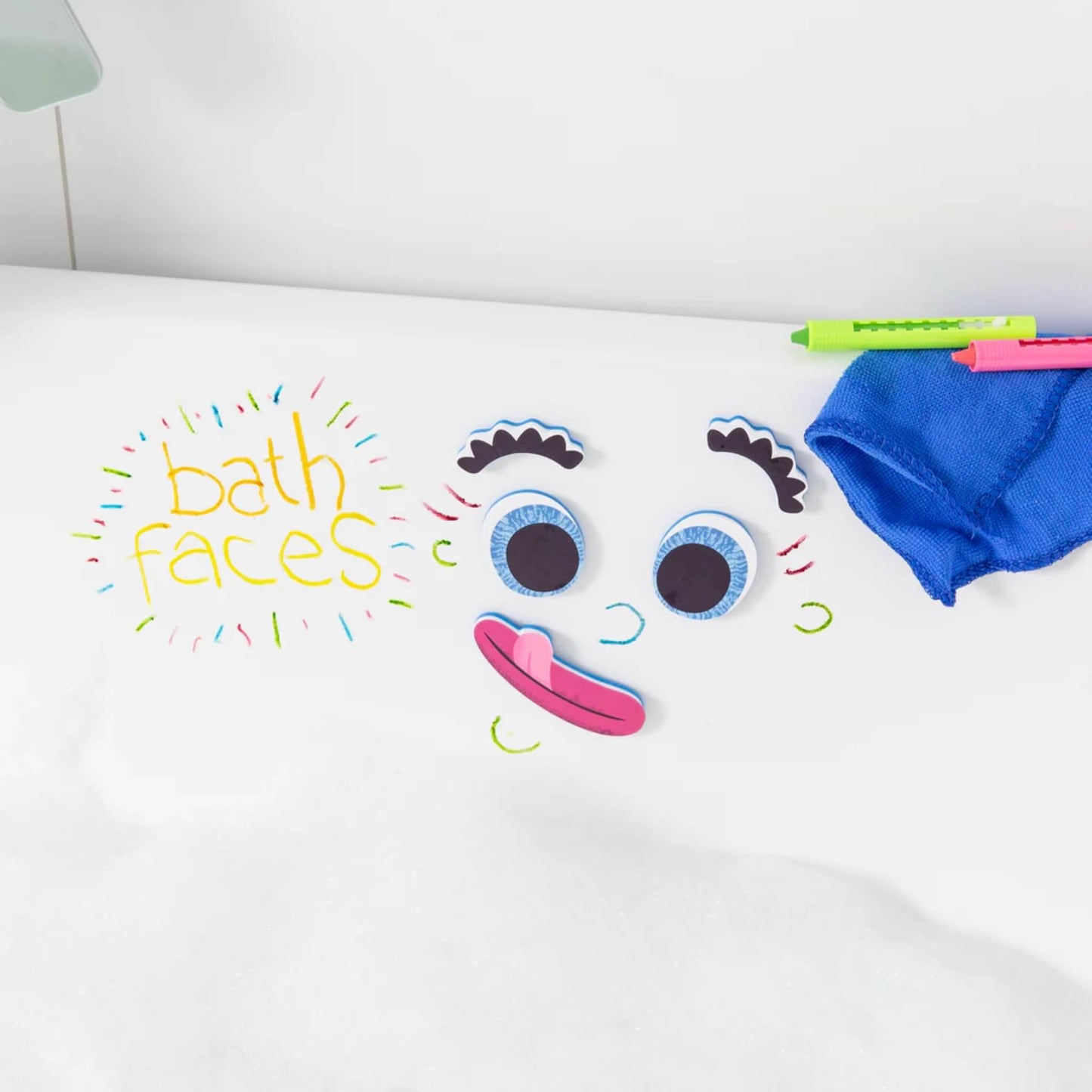Foam facial features and bath crayon drawings arranged on the side of a bathtub, with a blue cloth nearby.