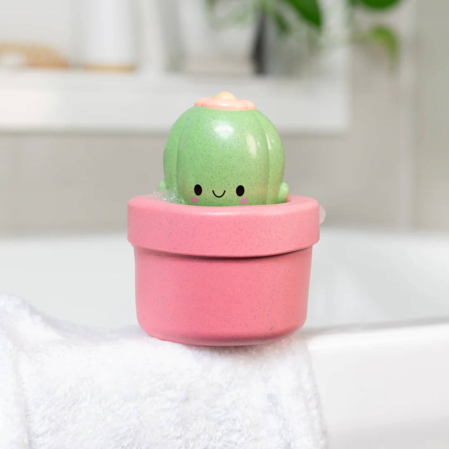 The cactus toy sitting on the edge of a white bathtub with bubbles and soft lighting.