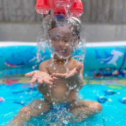 A toddler sitting in a paddling pool with the water cascading directly onto their head from the Bath Rocket above.