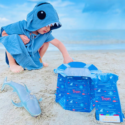 Child wearing a blue shark hooded towel crouches on sand beside an opened disposable potty, with a toy shark lying nearby.