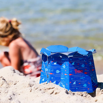 Blue disposable potty with fish design placed on sand at the beach, with a child blurred in the background near the shoreline.