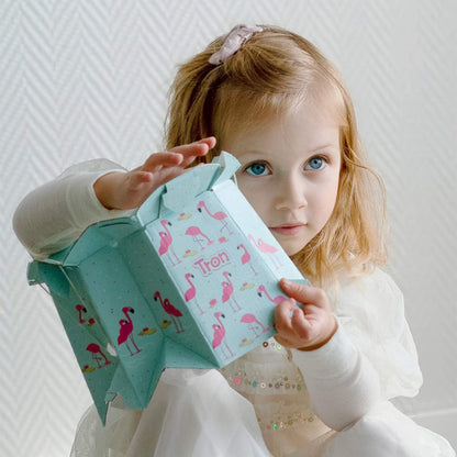 Young child with blonde hair holds a folded disposable potty in flamingo design, looking directly at the camera.