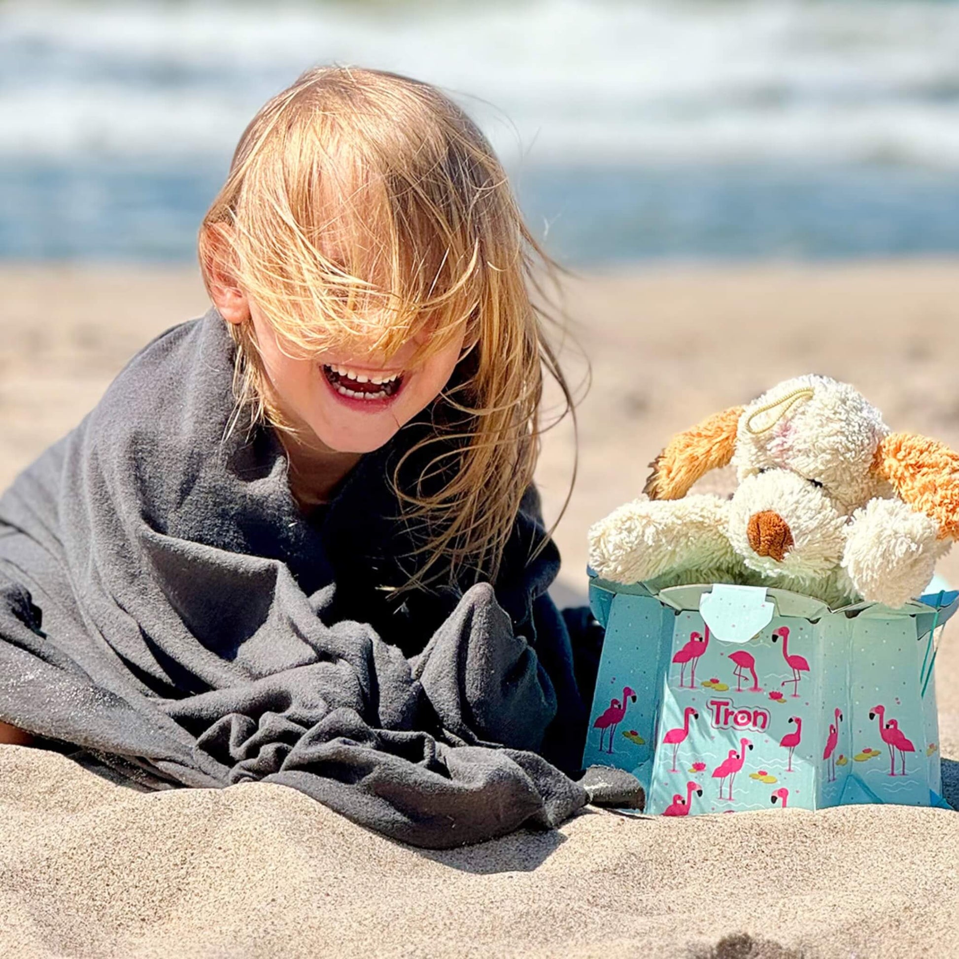 Smiling child wrapped in a blanket at the beach, sitting beside an assembled flamingo-print disposable potty with a soft toy placed inside.