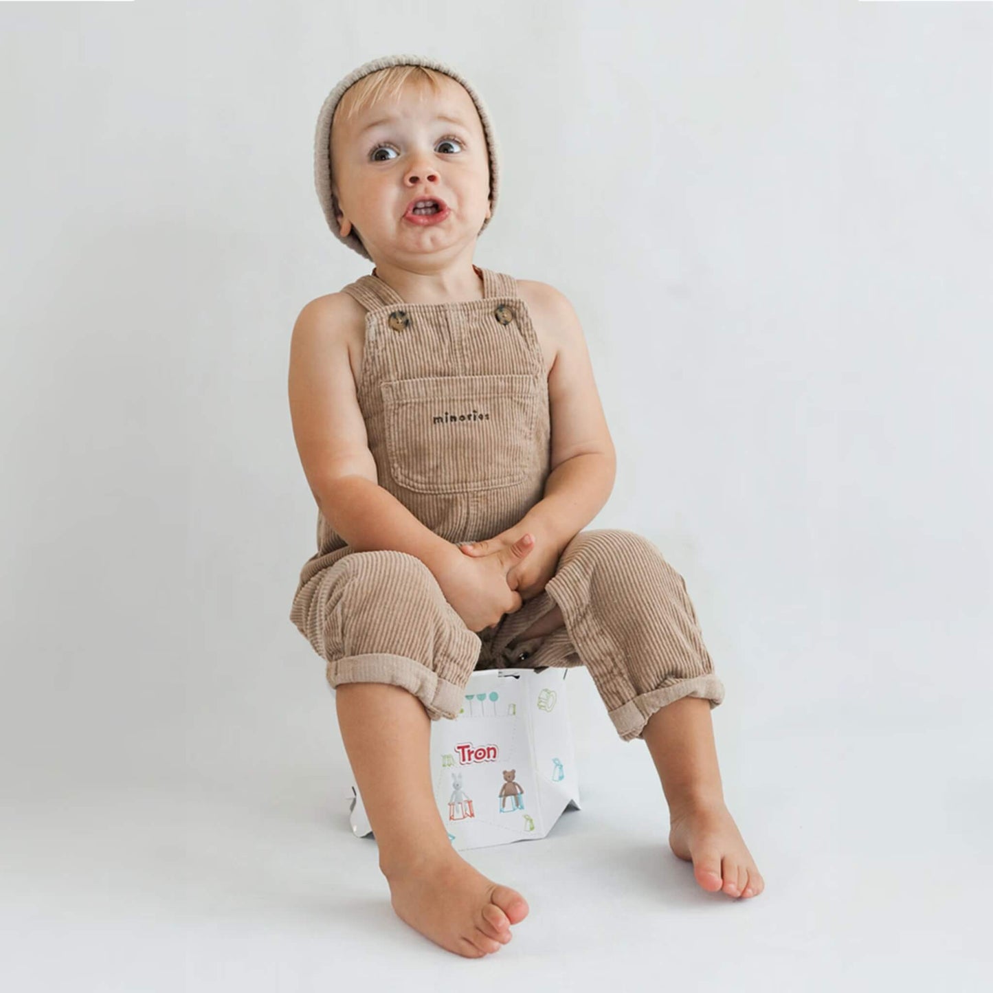 Toddler sitting on an open disposable potty, pulling a funny face while wearing cord dungarees.