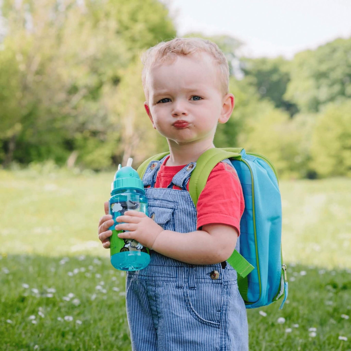 A toddler standing outdoors on a grassy field with trees in the background, wearing blue pinstriped overalls, a red t-shirt, a bright blue and green backpack, and holding a translucent teal water bottle.