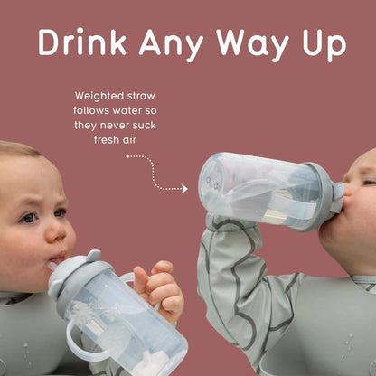 Two babies wearing grey bibs are shown drinking from clear plastic sippy cups with weighted straws against a dusty rose background. White text at the top reads 'Drink Any Way Up' with a dotted arrow pointing to the cup and smaller text explaining the weighted straw design.