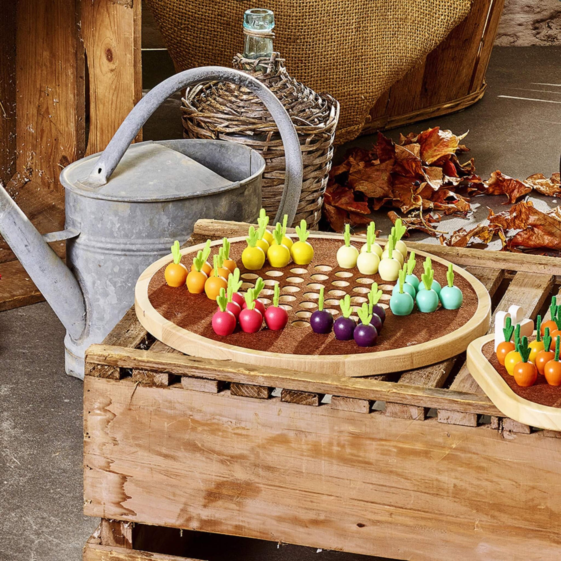 A wooden vegetable-themed board game displayed on a rustic wooden crate beside a metal watering can and wicker bottle in a cosy, autumnal setting.
