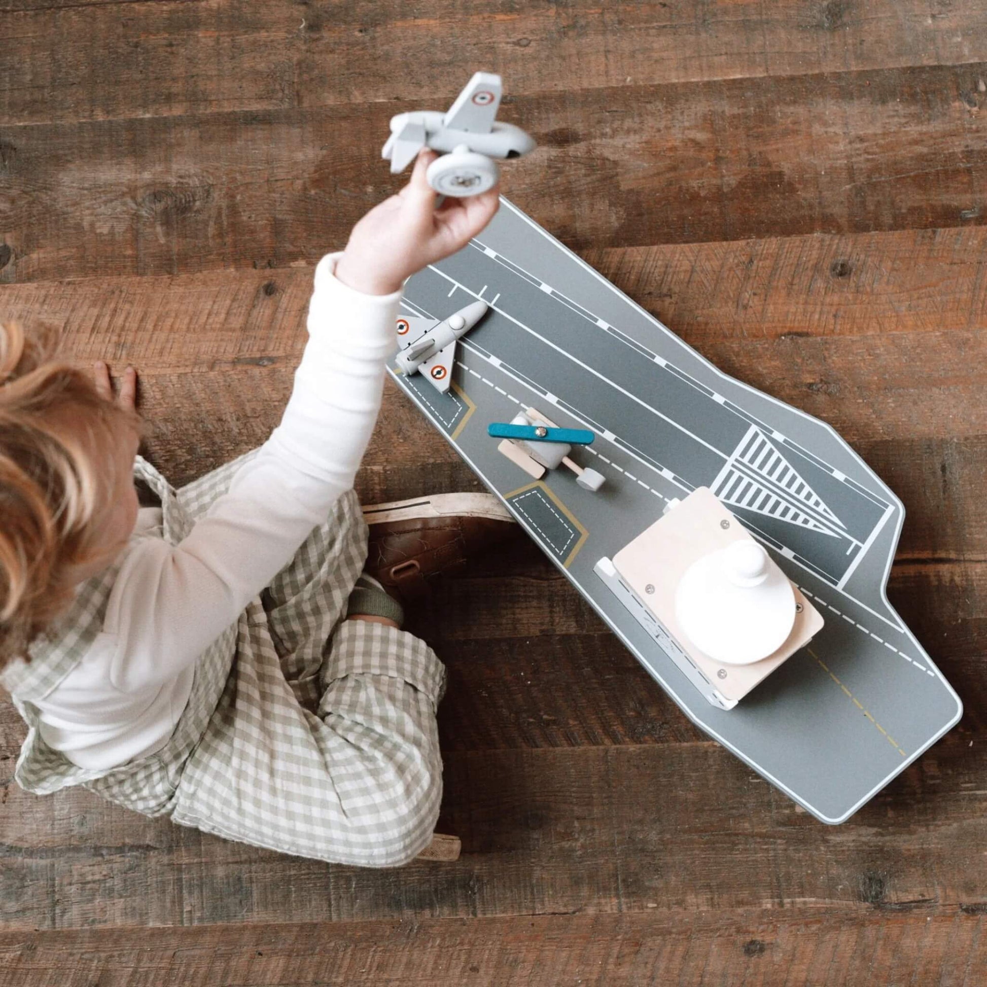 A young child sitting on a wooden floor holding a grey aircraft above a wooden aircraft carrier toy viewed from above.