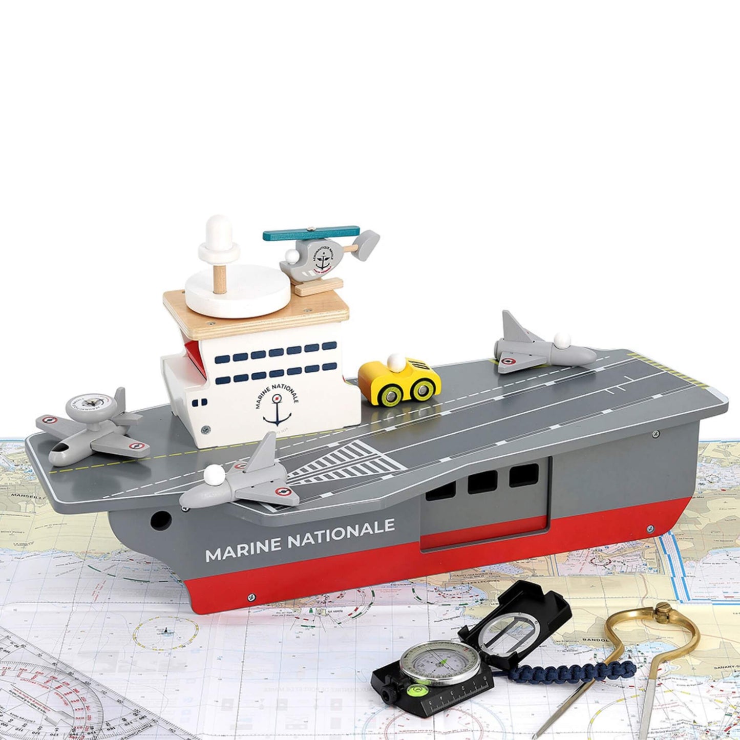 A wooden aircraft carrier toy positioned on nautical charts with navigation tools, showing three aircraft, a helicopter, and a yellow deck vehicle.