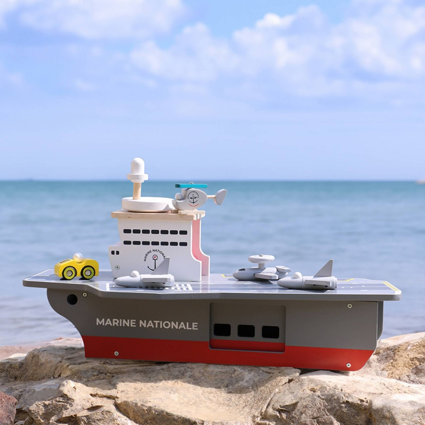A wooden aircraft carrier toy displayed outdoors on rocks with the sea in the background.