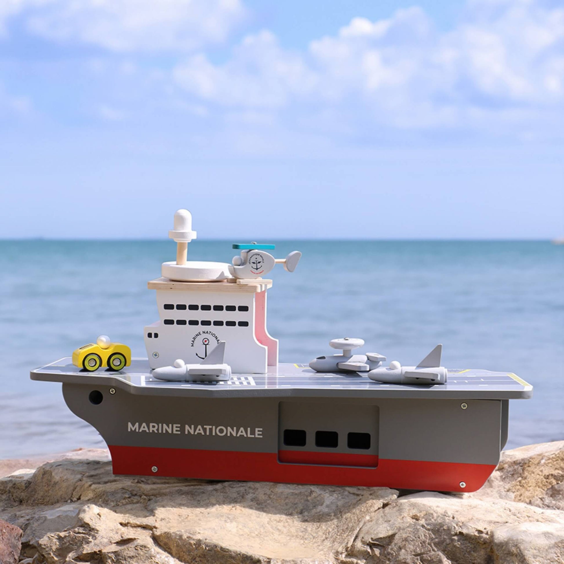 A wooden aircraft carrier toy displayed outdoors on rocks with the sea in the background.