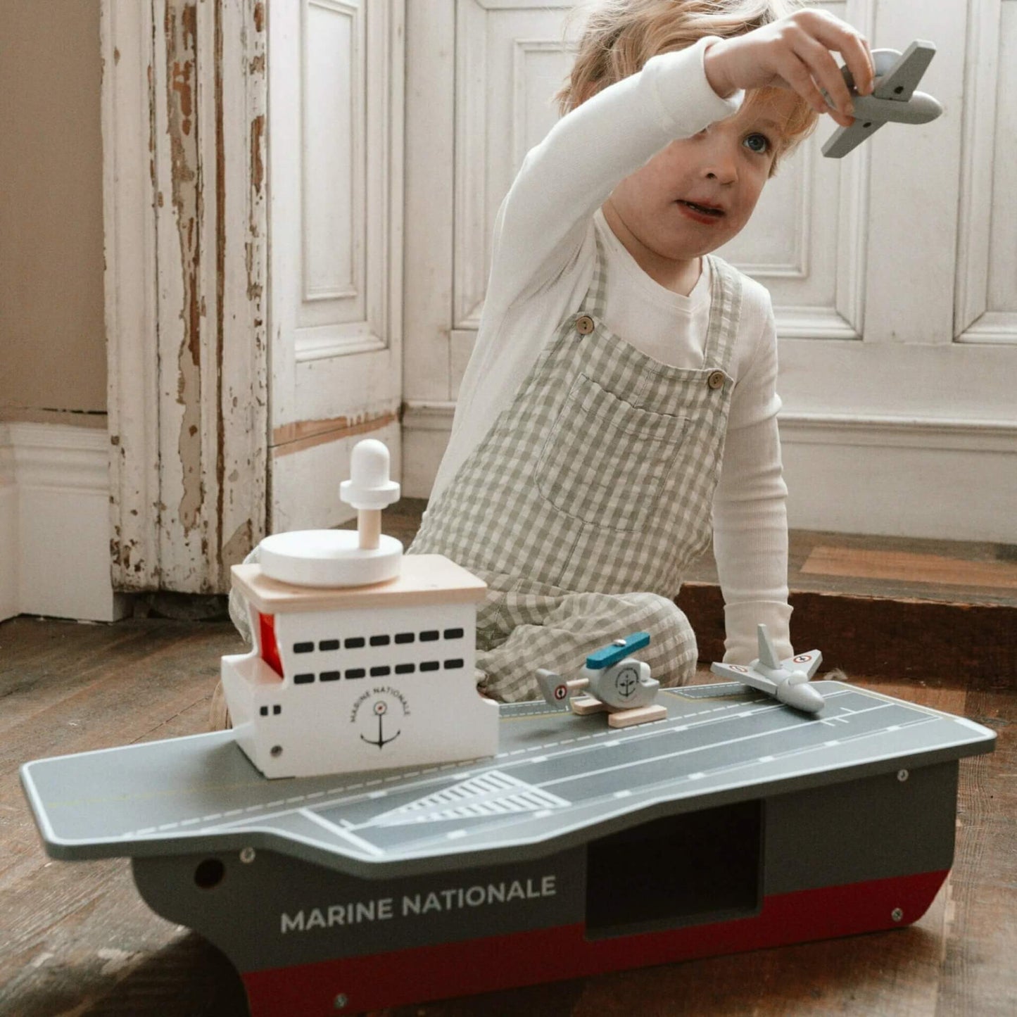 A young child playing indoors with a wooden aircraft carrier toy, holding a grey aircraft above the deck.