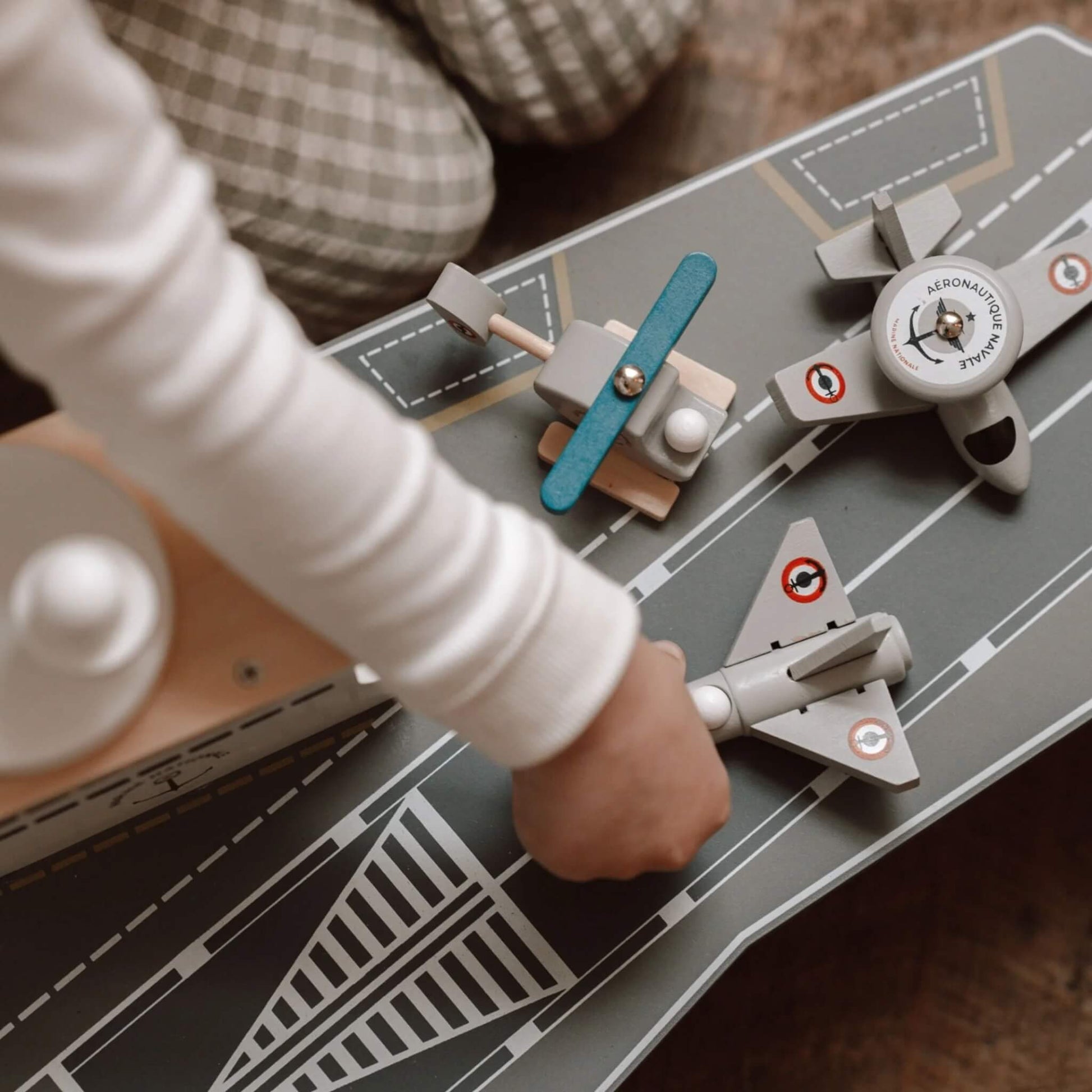 A close-up of a child’s hand placing a grey aircraft onto the deck of a wooden aircraft carrier toy alongside a helicopter and another aircraft.