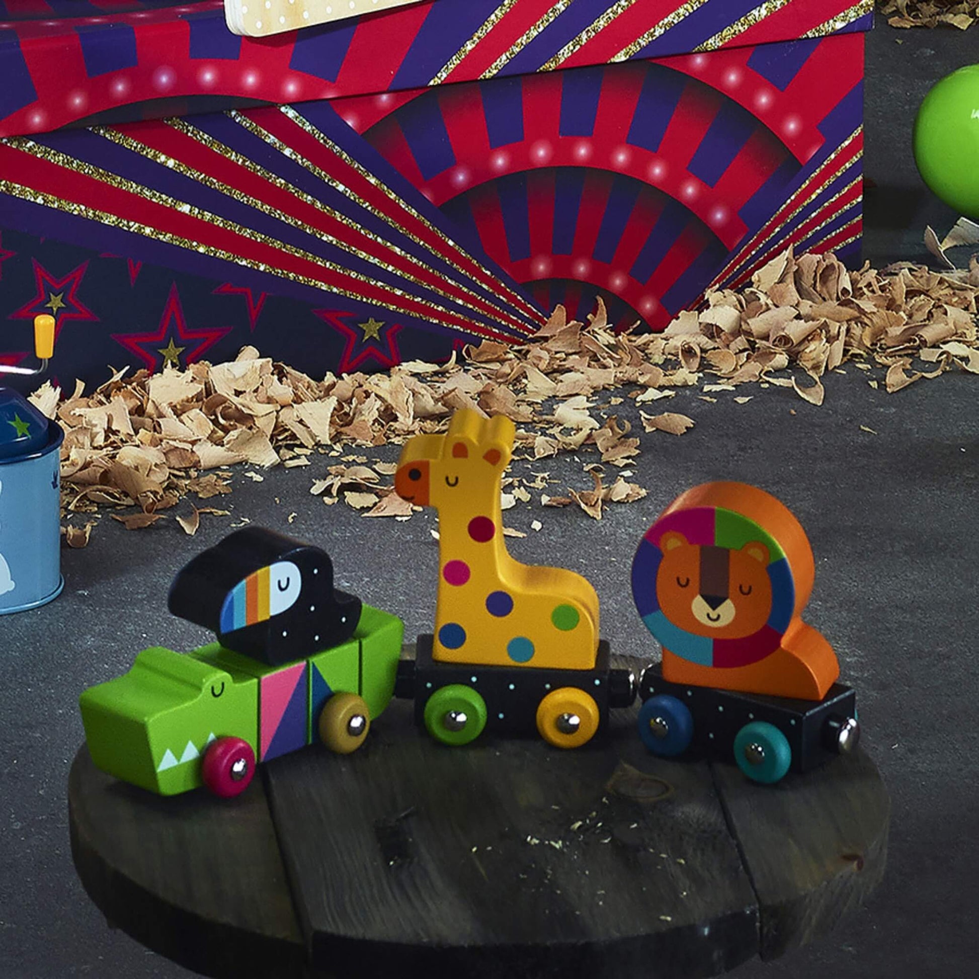 Three wooden animal train carriages—a crocodile with toucan, giraffe, and lion—placed on a round wooden table against a colourful circus-themed background.