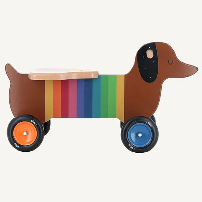 A wooden dachshund-shaped ride-on toy with rainbow stripe body, natural wood seat and four wheels in different bright colours.