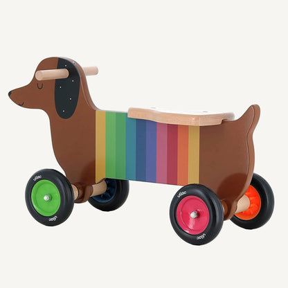A side profile of a wooden dog ride-on toy showing a long dachshund shape with rainbow stripes and smooth wooden seat.