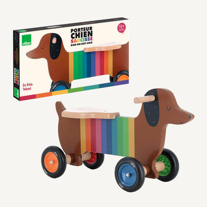 A boxed wooden dog ride-on toy with rainbow stripe design shown alongside the assembled ride-on, packaging displaying product illustration and branding.