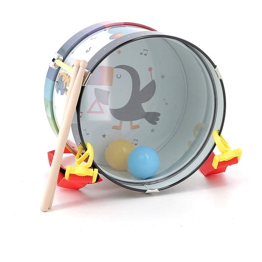 A metal toy drum lying on its side with a red strap, showing the inside decorated with a toucan holding a triangle and two plastic balls, alongside one wooden drumstick.