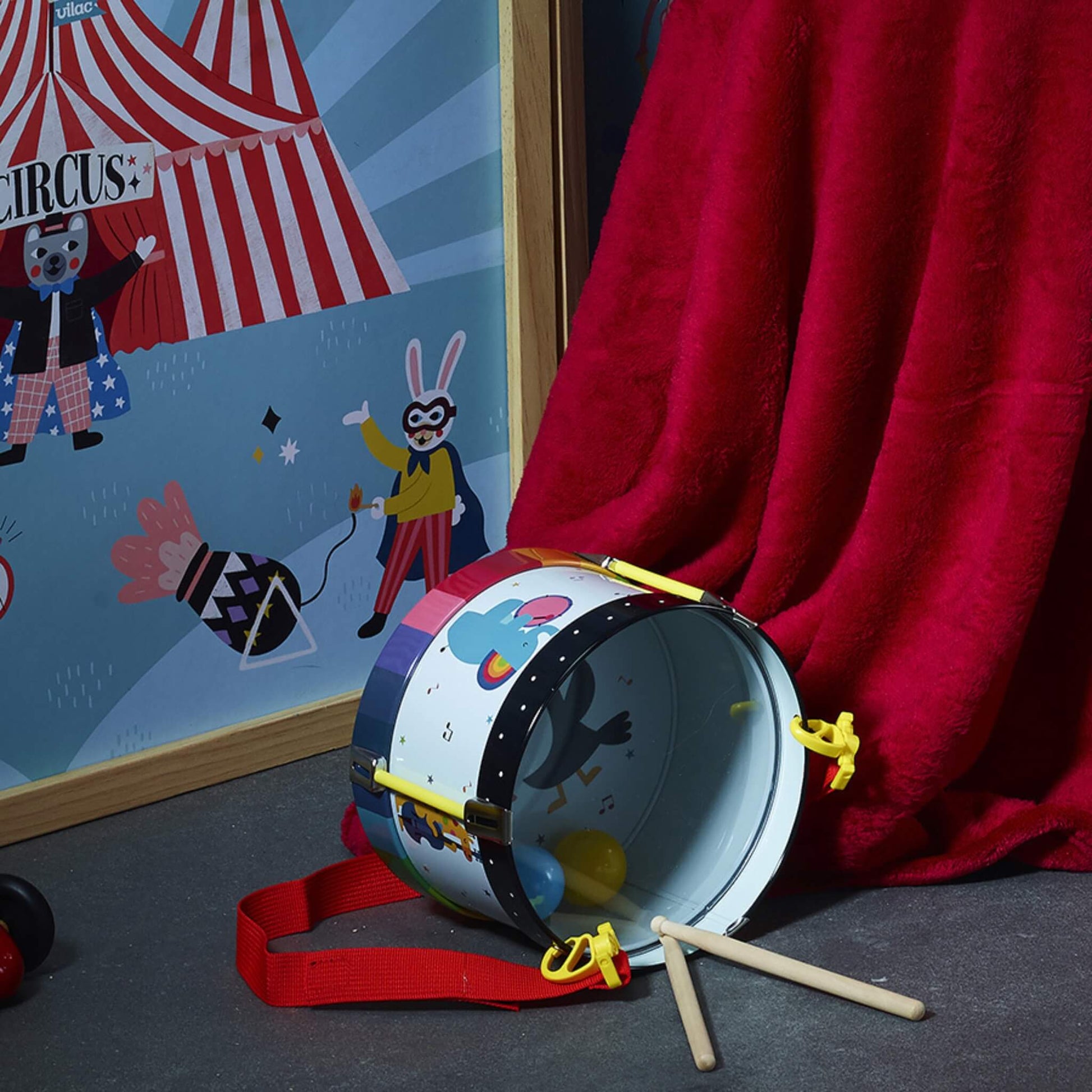 A metal toy drum lying on a floor next to a red curtain, with two wooden drumsticks and circus-themed wall art in the background.