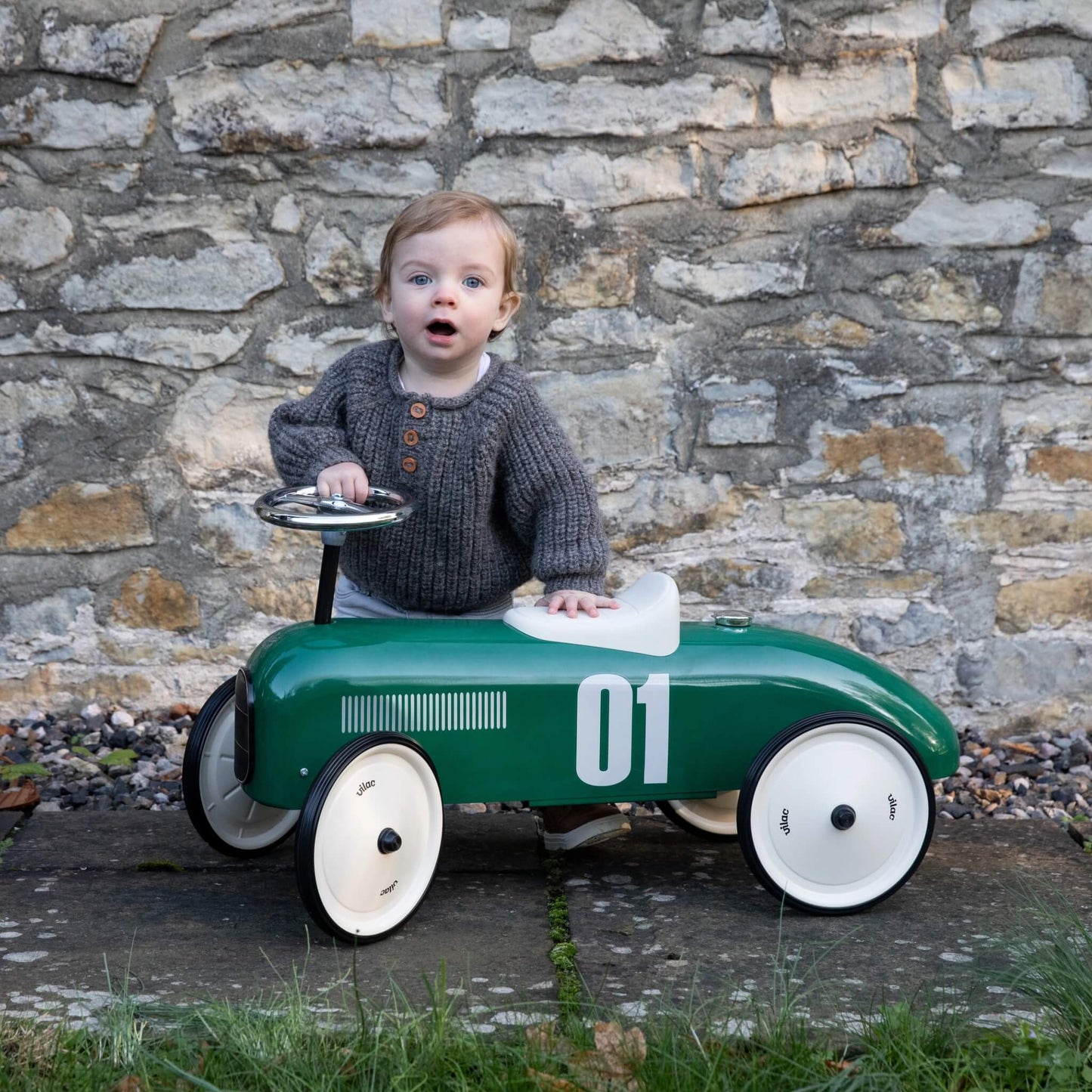 A young child standing beside a green ride-on toy car, holding the steering wheel while positioned next to a stone wall outdoors.